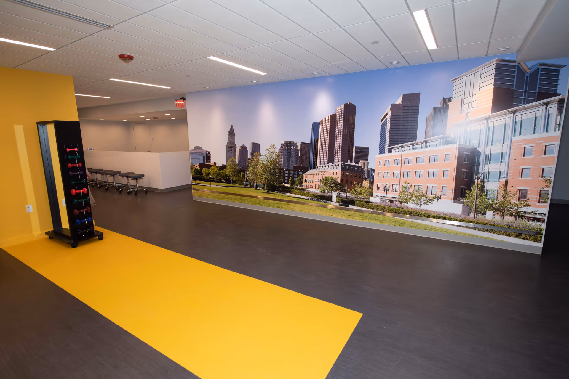 Spacious interior common area with a large city skyline wall mural, a yellow floor stripe, a rack of light dumbbells and stools.