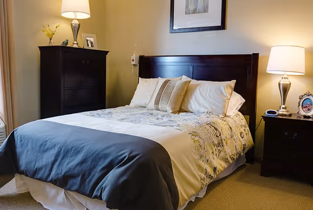 A neatly made bed with a dark wooden headboard, white and patterned bedding, and several pillows in a warmly lit bedroom. On the left side of the bed is a dark wooden dresser with a lamp, a small vase with yellow flowers, and a framed photo. On the right side is a matching nightstand with a lamp, a clock, and a framed picture. A framed artwork hangs on the wall above the bed.