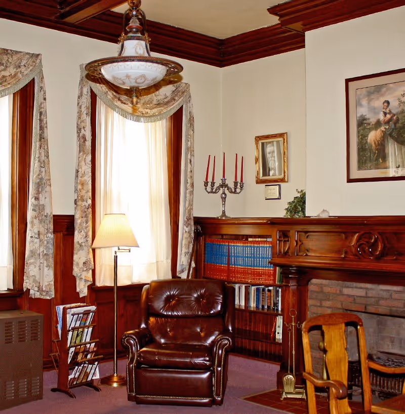 Traditional sitting room with a leather armchair in front of a fireplace, built-in bookshelves, a floor lamp, and draped windows.
