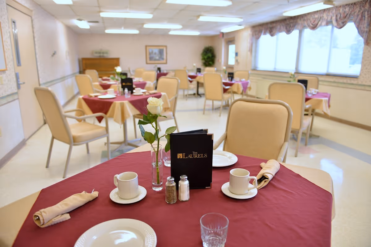 Dining room with round tables covered in burgundy tablecloths, place settings, a white rose centerpiece and a menu labeled "The Laurels".