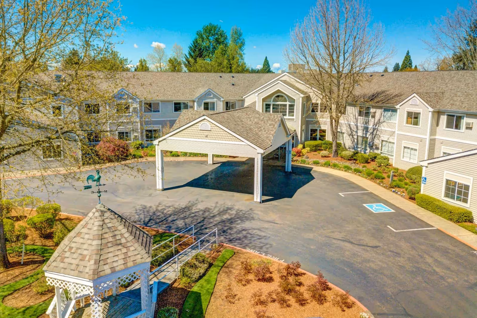 Exterior view of The Oaks at Lebanon senior living facility showing a two-story building with beige siding and multiple windows. There is a covered entrance driveway in the center and a small gazebo with a weather vane in the foreground. The surrounding area includes landscaped bushes, trees, and a clear blue sky.
