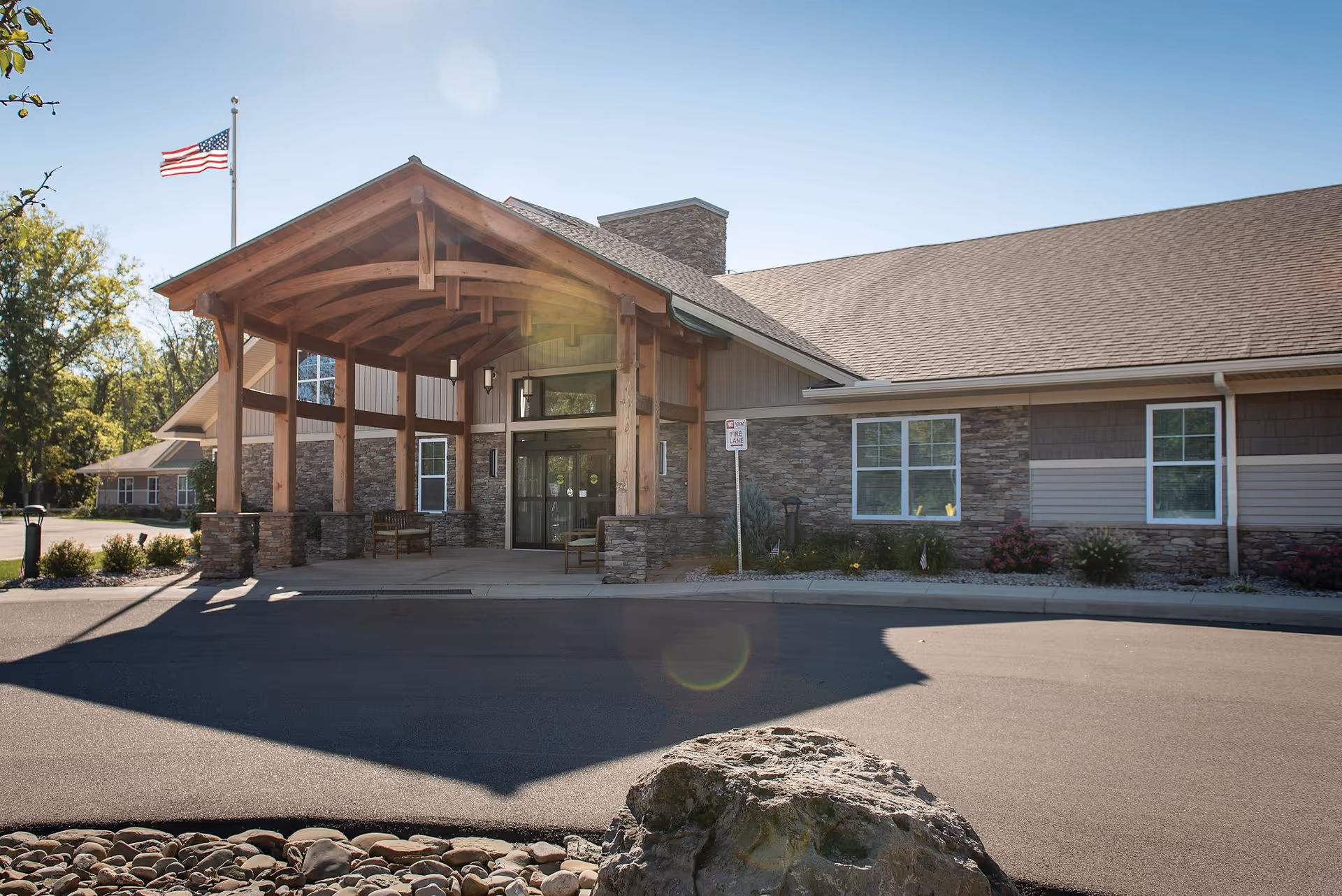 Exterior view of Legacy at Maiden Park facility entrance with a wooden covered porch, stone and siding walls, an American flag on a flagpole, and a clear blue sky.