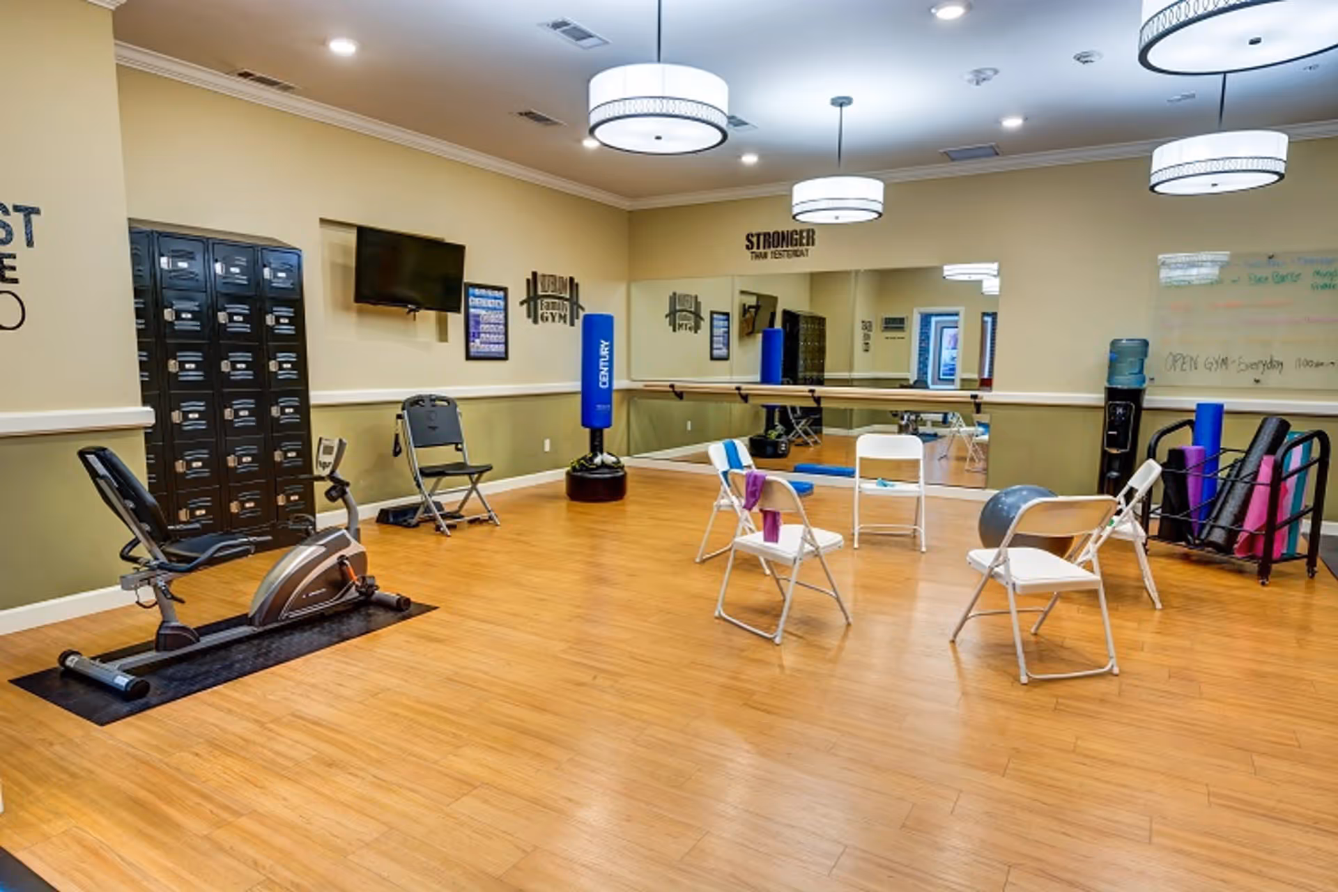 A fitness room with wooden flooring featuring exercise equipment including a recumbent bike, a punching bag, and various yoga mats and exercise balls. Several white folding chairs are arranged in a circle in the center of the room. The walls have mirrors, motivational text, and a mounted TV. Lockers are visible along one wall.
