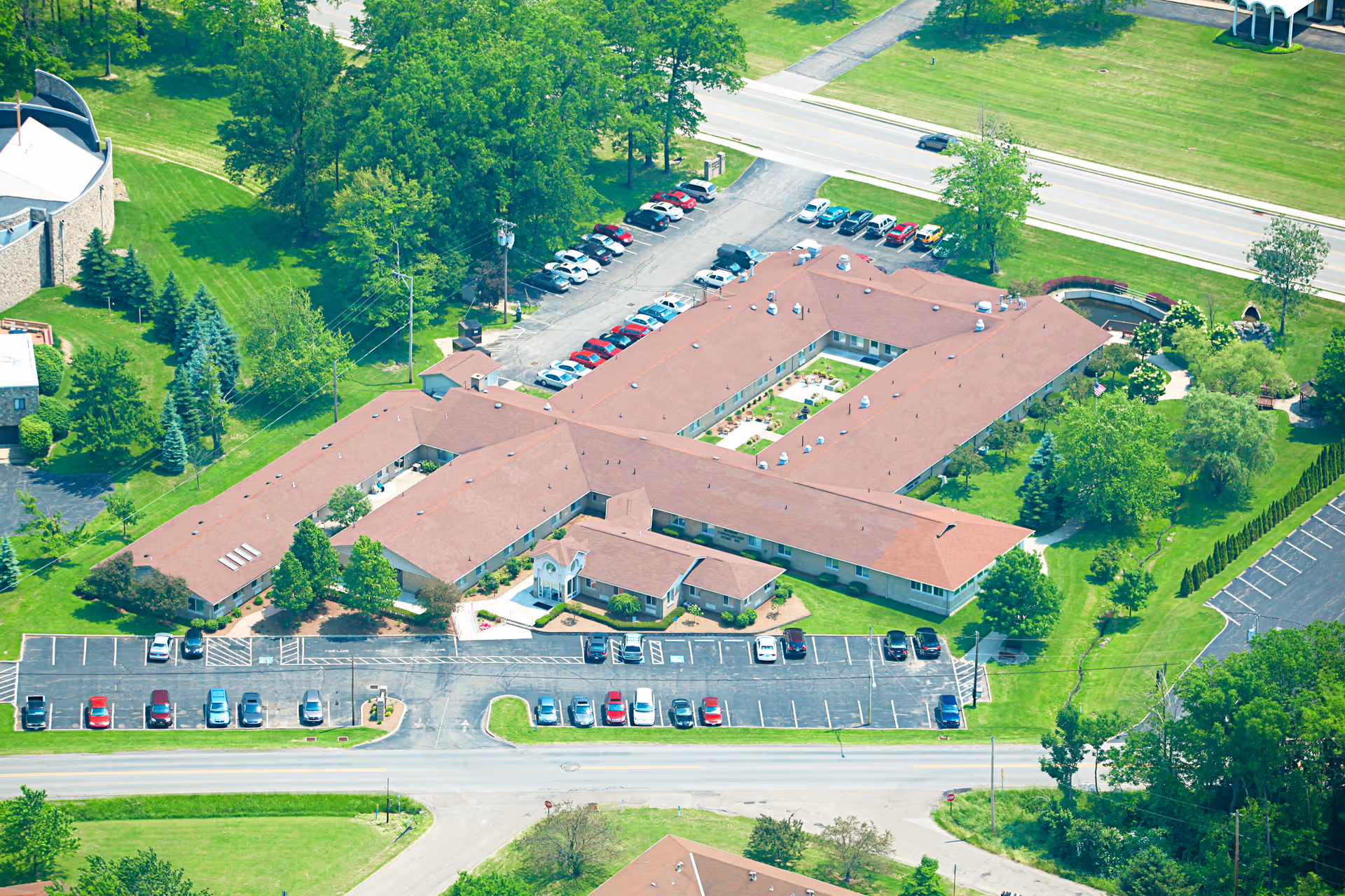 Aerial view of a senior living facility named Hermitage, showing a large building with a red roof surrounded by green lawns, trees, and parking lots with several cars parked. Roads border the property on two sides.