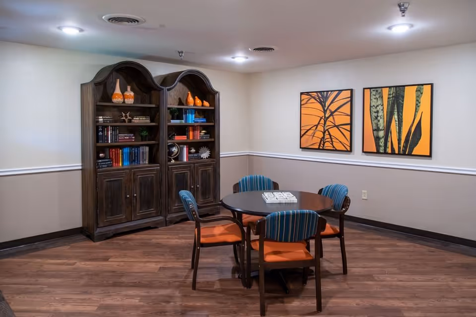 A cozy interior room with a round table surrounded by four chairs with blue and orange upholstery. Behind the table is a large wooden bookshelf filled with books and decorative items. Two framed artworks featuring abstract plant designs in orange and black hang on the wall. The room has wood flooring and soft overhead lighting.