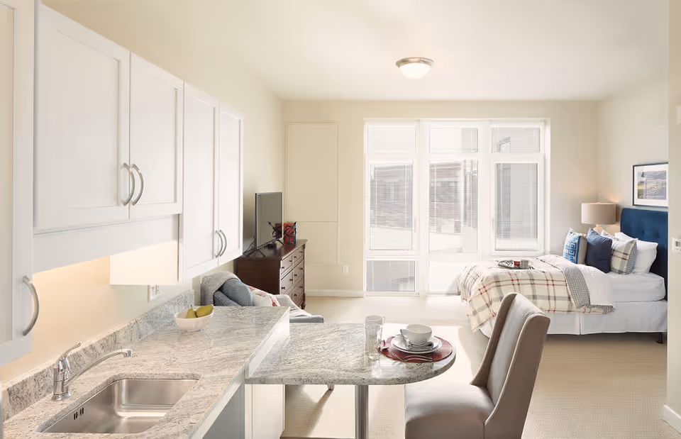 Bright and airy studio apartment with a small kitchen area featuring white cabinets and a marble countertop with a sink. A dining table with two place settings is adjacent to the kitchen. The living and sleeping area includes a bed with plaid bedding, a blue upholstered headboard, a wooden dresser with a TV, and large windows letting in natural light.