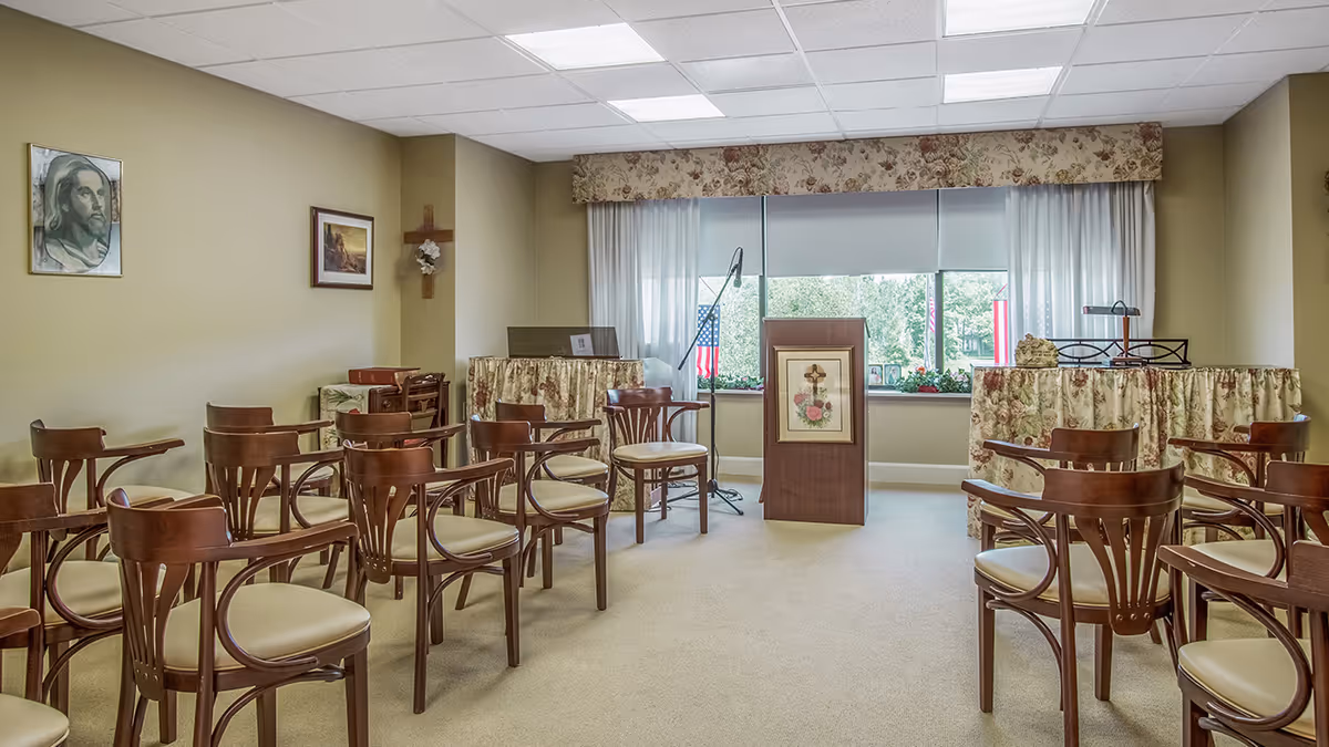 A small chapel or prayer room with rows of wooden chairs with cream cushions facing a wooden podium adorned with a floral cross picture. The room has beige walls decorated with religious artwork, including a portrait of Jesus and a wooden cross with a flower. There is a large window at the back with floral valances and sheer white curtains, letting in natural light and showing greenery outside. Two tables covered with floral fabric are positioned on either side of the podium, one with a microphone stand and the other with a lamp and decorative items.