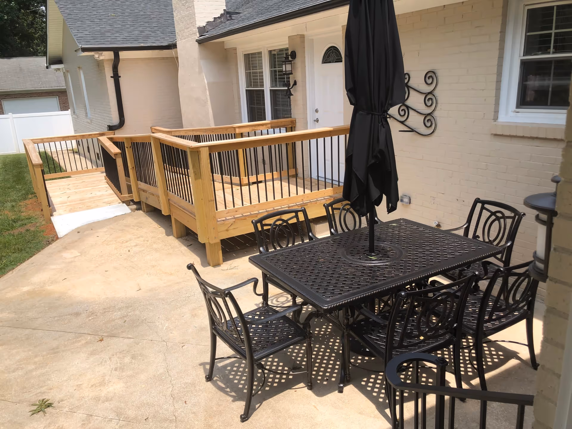 Outdoor patio area with a black metal table and six matching chairs. The table has a closed black umbrella in the center. Behind the patio is a wooden ramp with black metal railings leading to a white door on a light-colored brick building.
