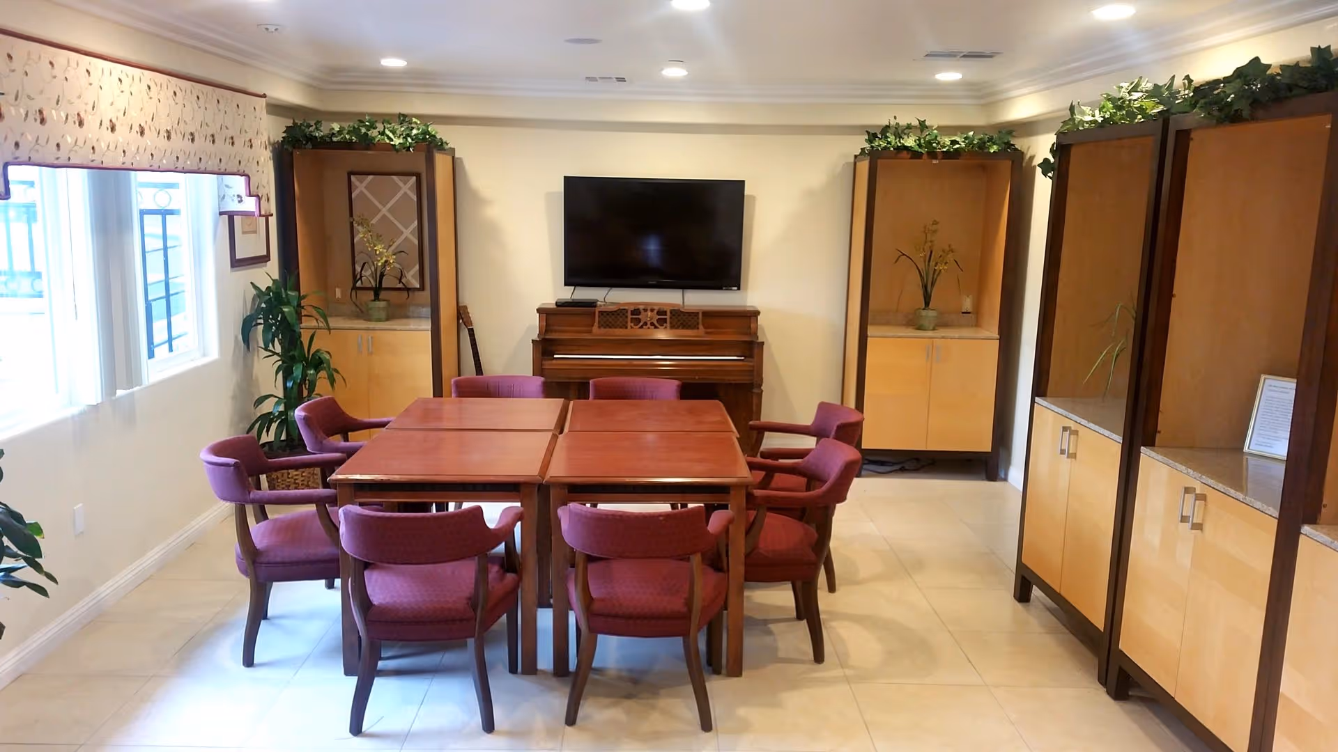A well-lit room with four wooden tables pushed together surrounded by eight purple cushioned chairs. A piano with a flat-screen TV mounted above it is against the far wall. The room has light-colored tiled flooring, built-in wooden cabinets with plants on top, and a window with a floral valance on the left side.