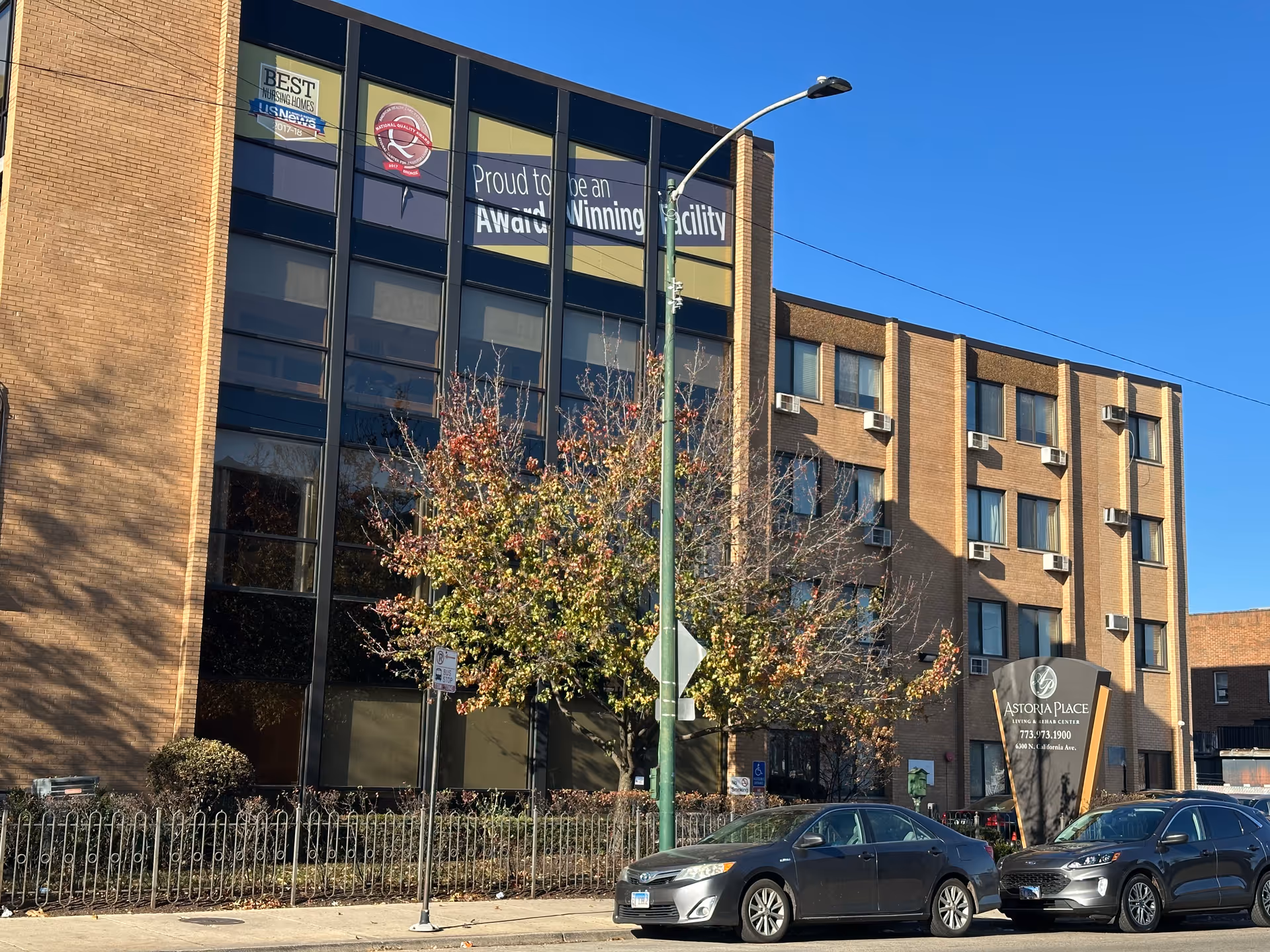 Front exterior of a multi-story brick senior living building with an Astoria Place sign, parked cars, and a tree under a clear blue sky.