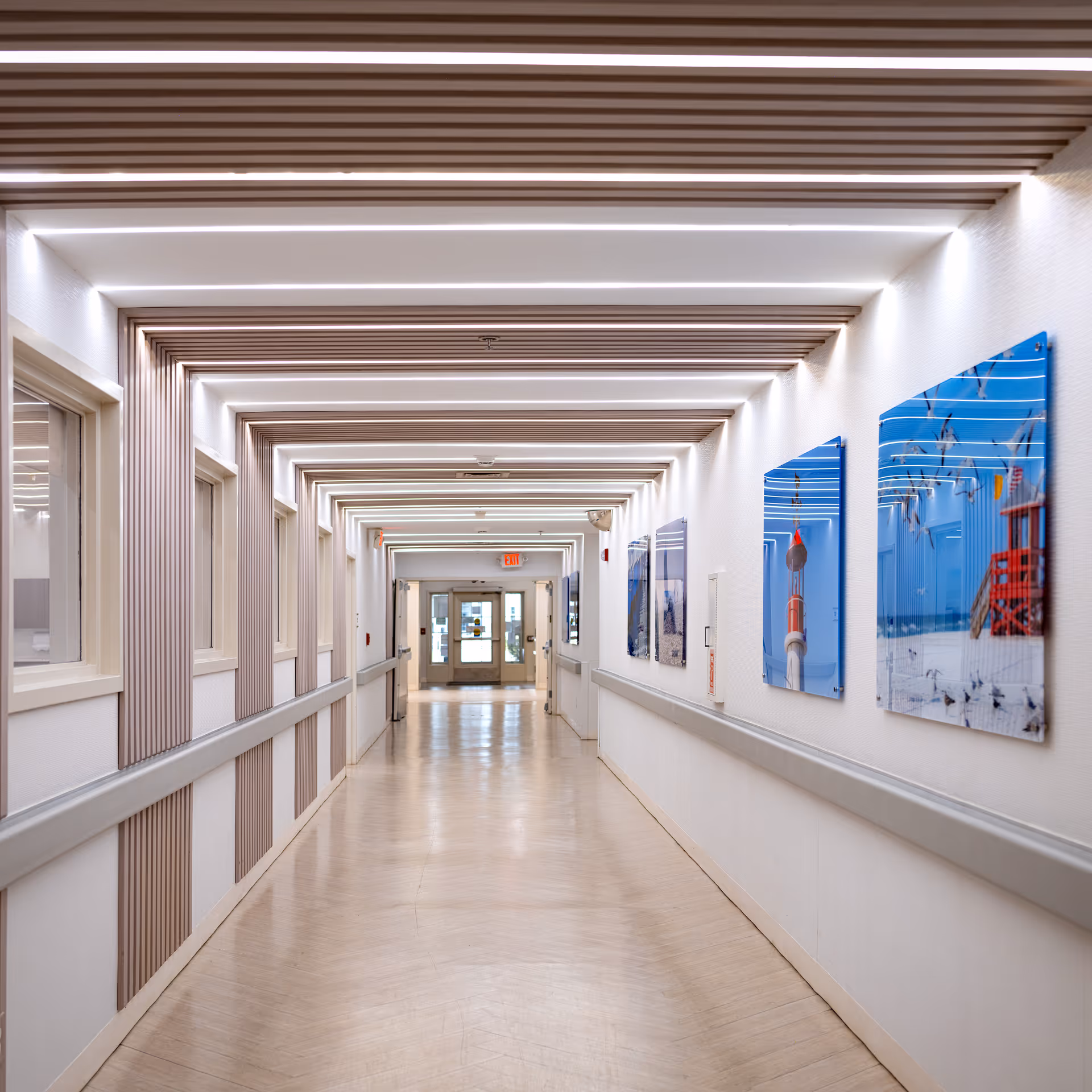 A long, well-lit hallway with modern ceiling light fixtures and wooden panel accents on the walls. The hallway features handrails on both sides and several framed pictures of beach scenes on the right wall. At the end of the hallway, there is a glass door with an exit sign above it.