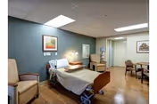 A well-lit hospital-style room with a single adjustable bed, two beige armchairs, a small table with chairs in the background, and framed artwork on the walls. The floor is wooden, and the walls are painted in muted tones.