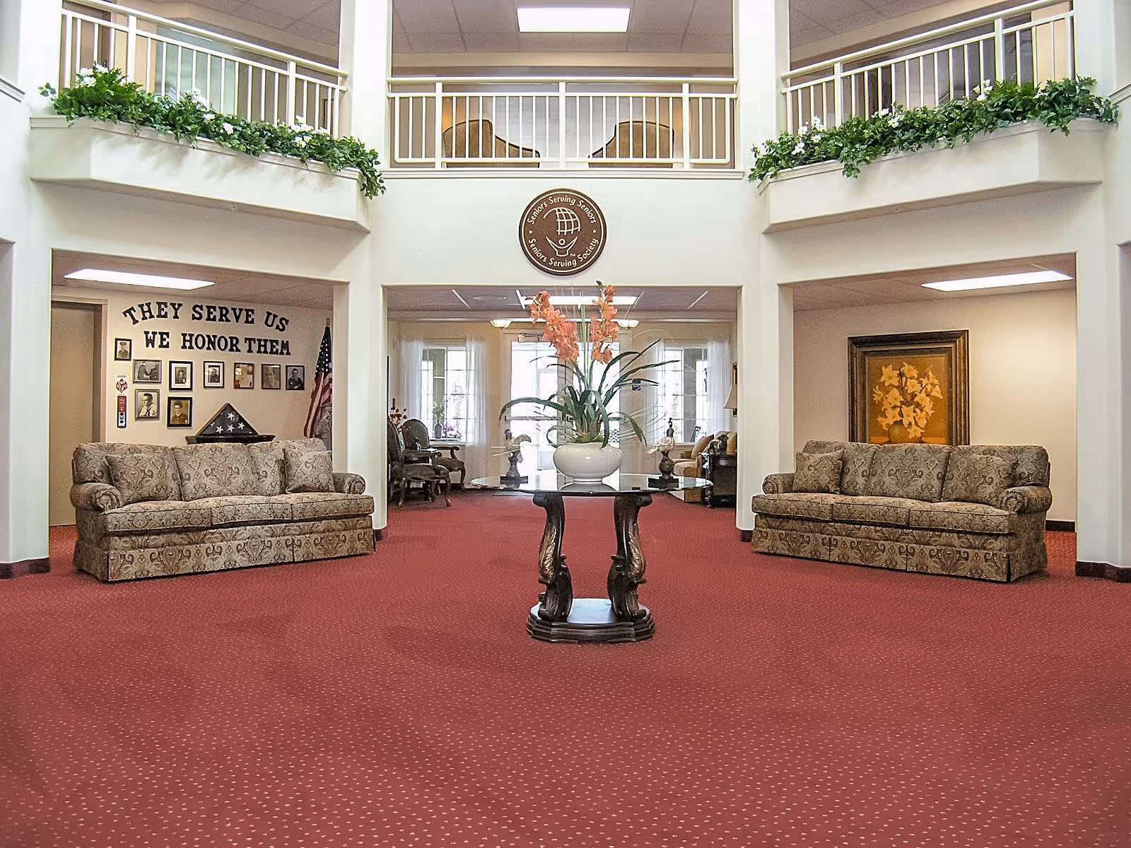 A spacious senior living facility lobby with red carpet flooring, two patterned sofas facing each other, a round wooden table with a large floral arrangement in the center, and a second-floor balcony with plants along the railing. On the left wall, there is a memorial display with the text 'THEY SERVE US WE HONOR THEM' and framed photos, along with an American flag. The right wall features a framed floral painting.