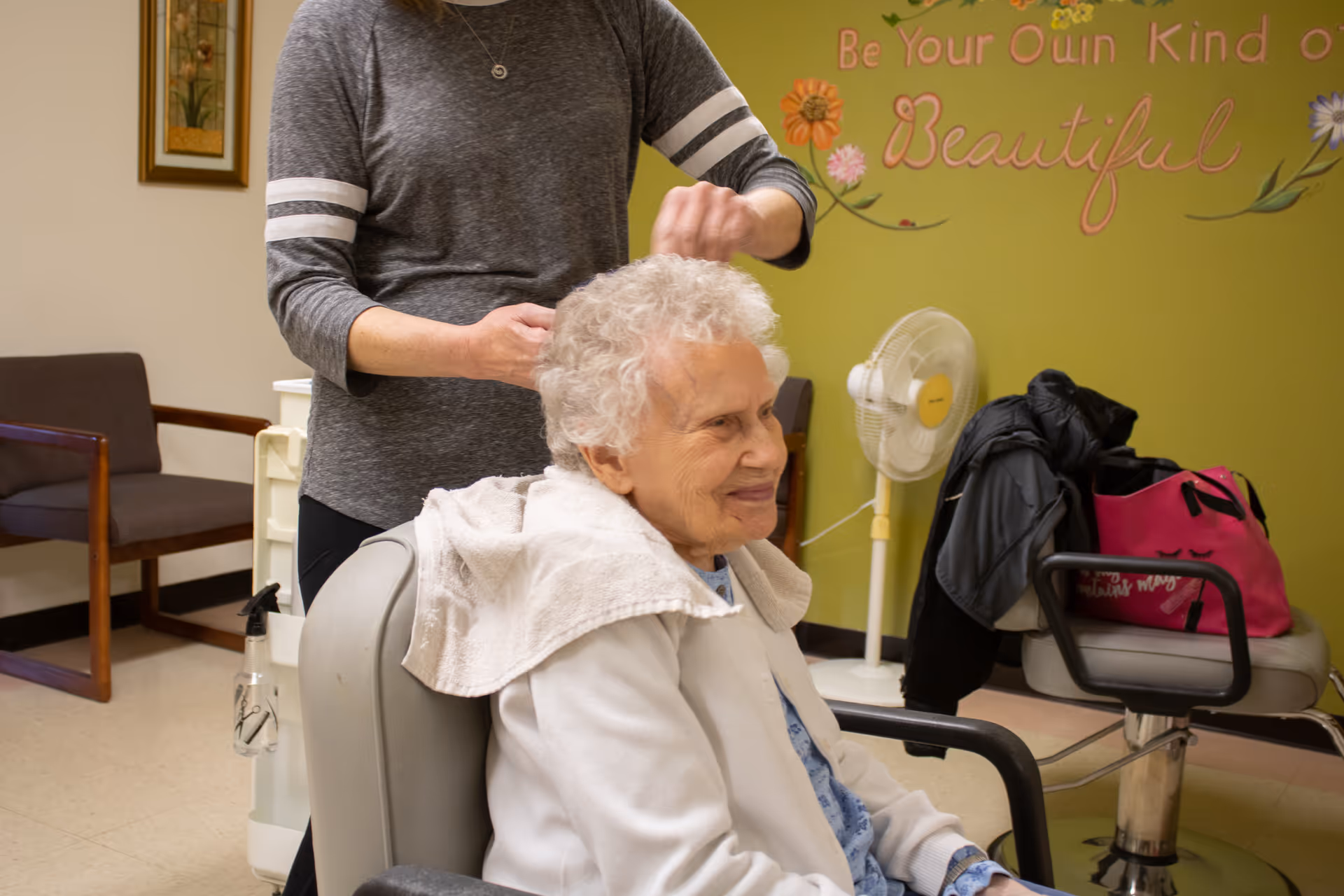 An elderly woman with white curly hair sits in a salon chair with a towel draped over her shoulders while a person standing behind her styles her hair. The room has a green wall with floral decorations and the words 'Be Your Own Kind of Beautiful' written on it. There is a fan, a chair with a black jacket and a pink bag on it, and a framed picture on the adjacent wall.
