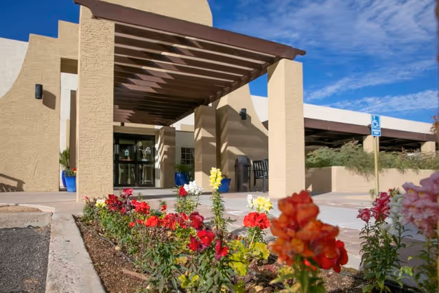 Entrance of Shea Post Acute Rehabilitation Center with a beige stucco building, a wooden pergola over the walkway, colorful flowers planted along the path, and a clear blue sky above.