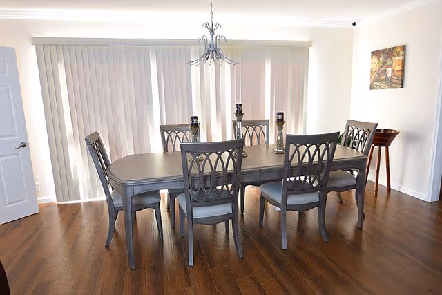 A dining room with a rectangular wooden dining table surrounded by six matching chairs with cushioned seats. The room has wooden flooring, vertical blinds covering large windows, a chandelier hanging above the table, and a small side table with a painting on the wall.