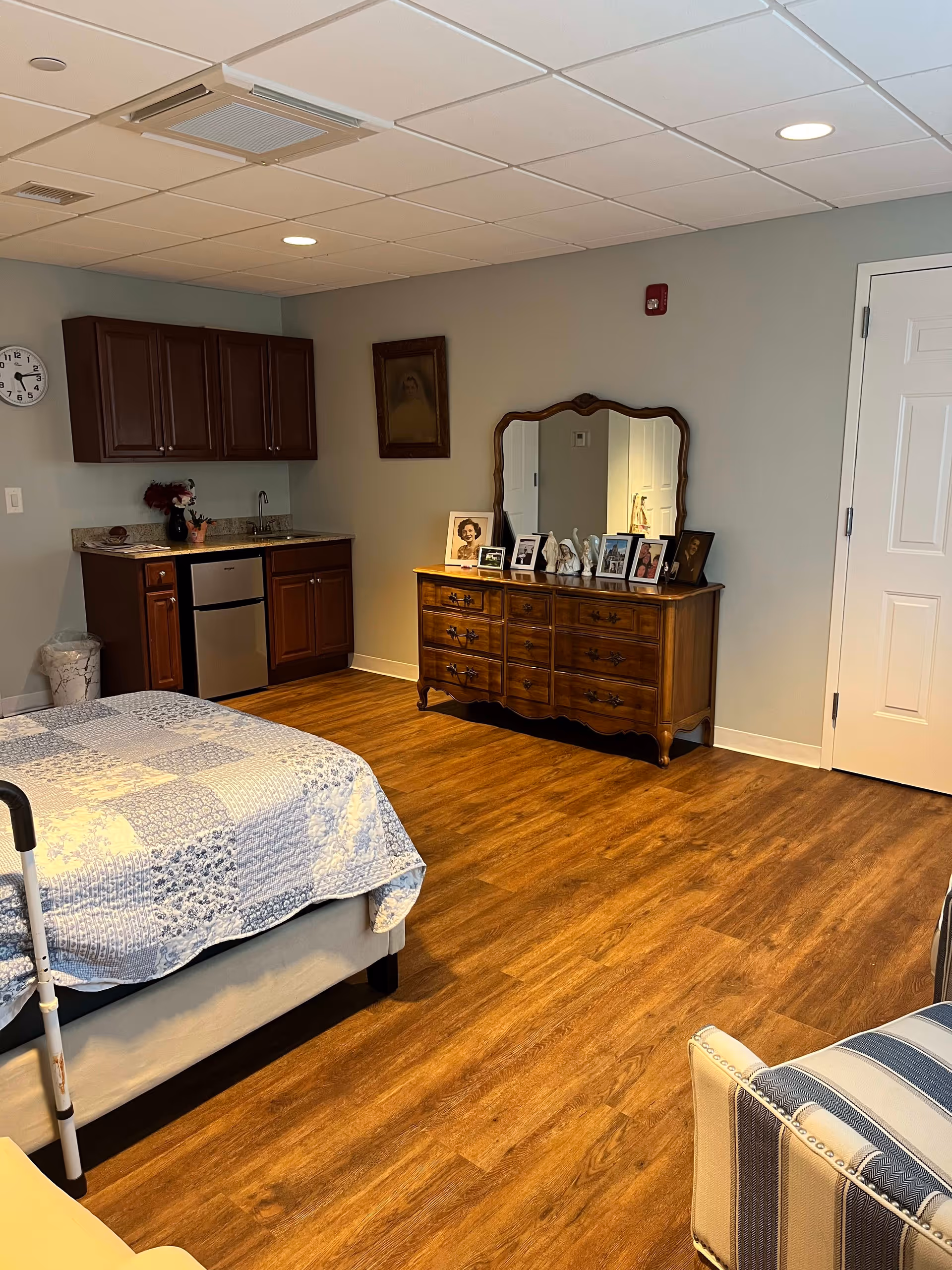 A senior living facility room with a bed covered in a blue and white patchwork quilt, a wooden dresser with a large mirror and several framed photos on top, a small kitchenette with dark wood cabinets, a sink, and a mini dishwasher, and a striped upholstered chair in the foreground. The room has wood flooring and light gray walls.