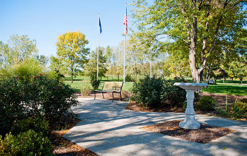 Outdoor garden area with a concrete pathway, a wooden bench, a white birdbath, green bushes, trees, and two flagpoles with flags under a clear blue sky.