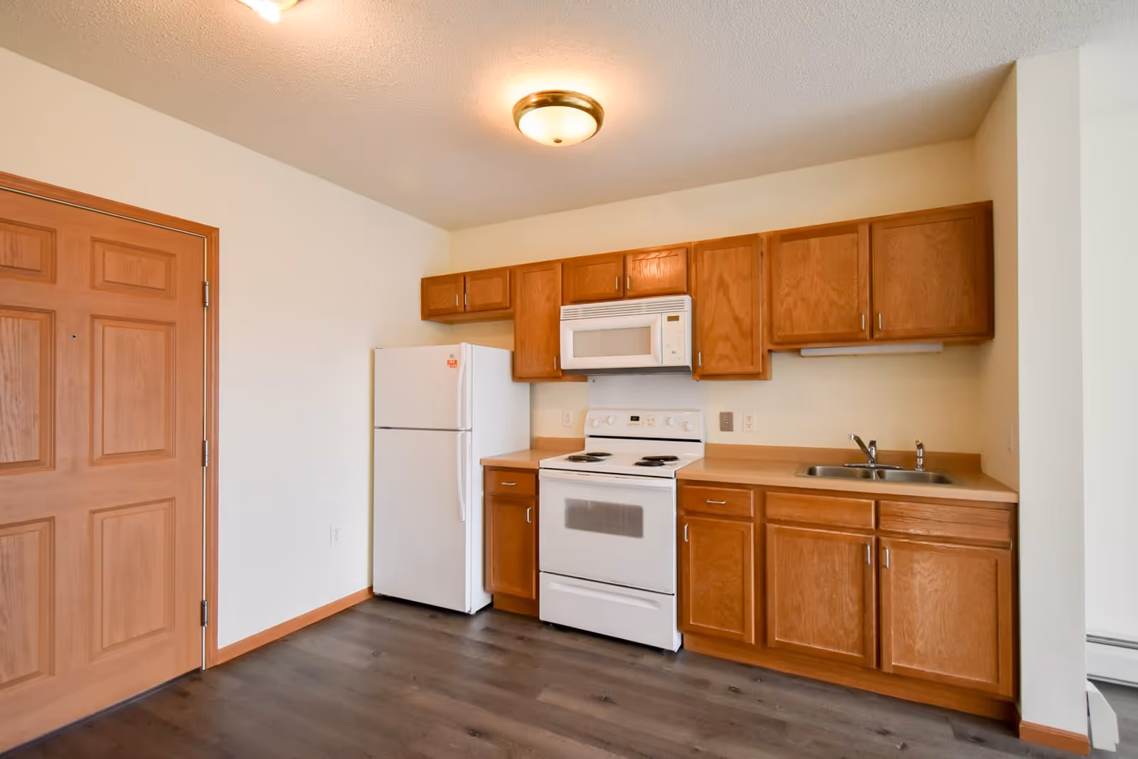 A small kitchen area in a senior apartment featuring wooden cabinets, a white refrigerator, a white electric stove with an oven, a white microwave above the stove, a stainless steel sink, and a wooden door to the left. The floor is dark wood, and the walls are painted white.