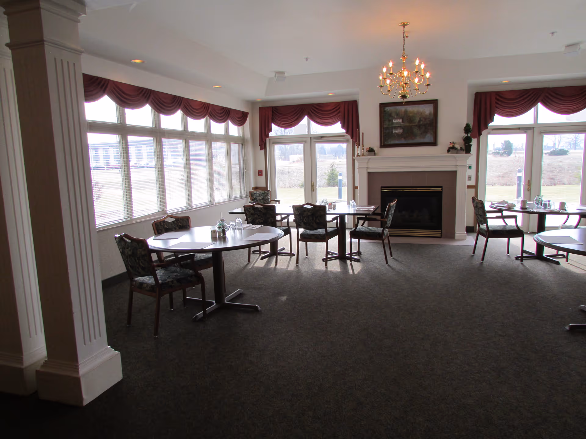 A dining area with round tables and chairs arranged around them. The room has large windows with maroon valances, allowing natural light to fill the space. There is a fireplace with a painting above it and a chandelier hanging from the ceiling. The carpet is dark-colored and the walls are light-colored.