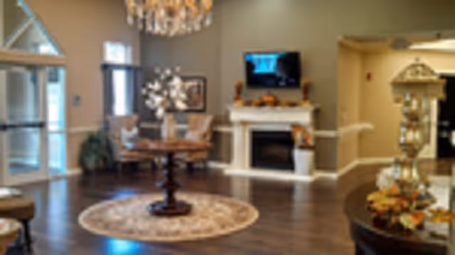A cozy living room area with a round wooden table on a circular rug in the center, surrounded by upholstered chairs. There is a fireplace with a TV mounted above it on the far wall, and a chandelier hanging from the ceiling. The room has wooden flooring and neutral-colored walls, with large windows and a glass door letting in natural light.
