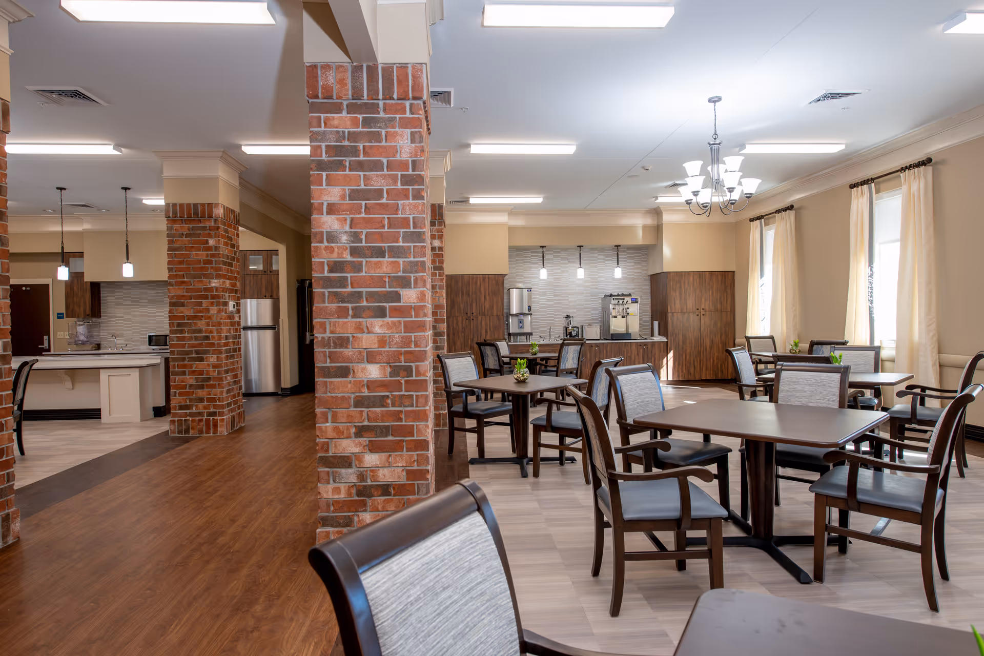 A spacious dining area in an assisted living facility featuring multiple wooden tables and chairs with cushioned seats. The room has large windows with cream-colored curtains allowing natural light to fill the space. There are brick pillars and a kitchen area in the background with stainless steel appliances and pendant lighting.