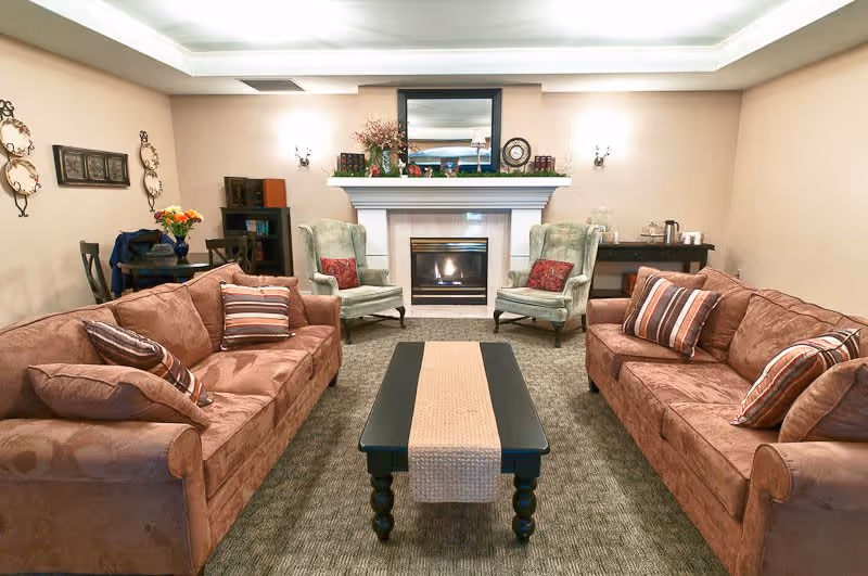 A cozy living room with two brown sofas facing each other, each with striped and solid pillows. Between them is a black coffee table with a beige runner. At the far end of the room is a white fireplace with a fire burning, flanked by two green armchairs with red pillows. Above the fireplace is a large mirror and decorative items. The walls are beige with wall sconces and framed decorations. There is a small table with flowers and chairs on the left side and a sideboard with a coffee setup on the right.