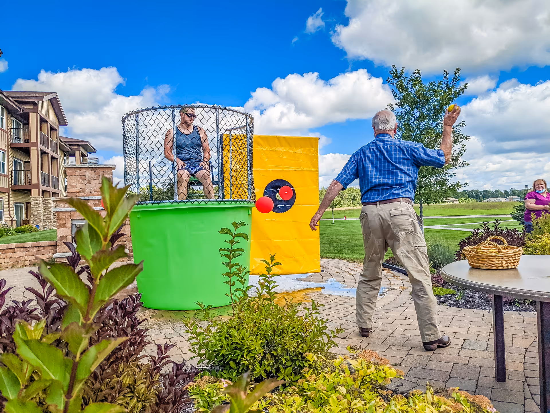 An elderly man throws a ball at a dunk tank outdoors while a woman sits inside the dunk tank cage. Another woman watches nearby, and the scene is set in a garden area with a building and blue sky with clouds in the background.