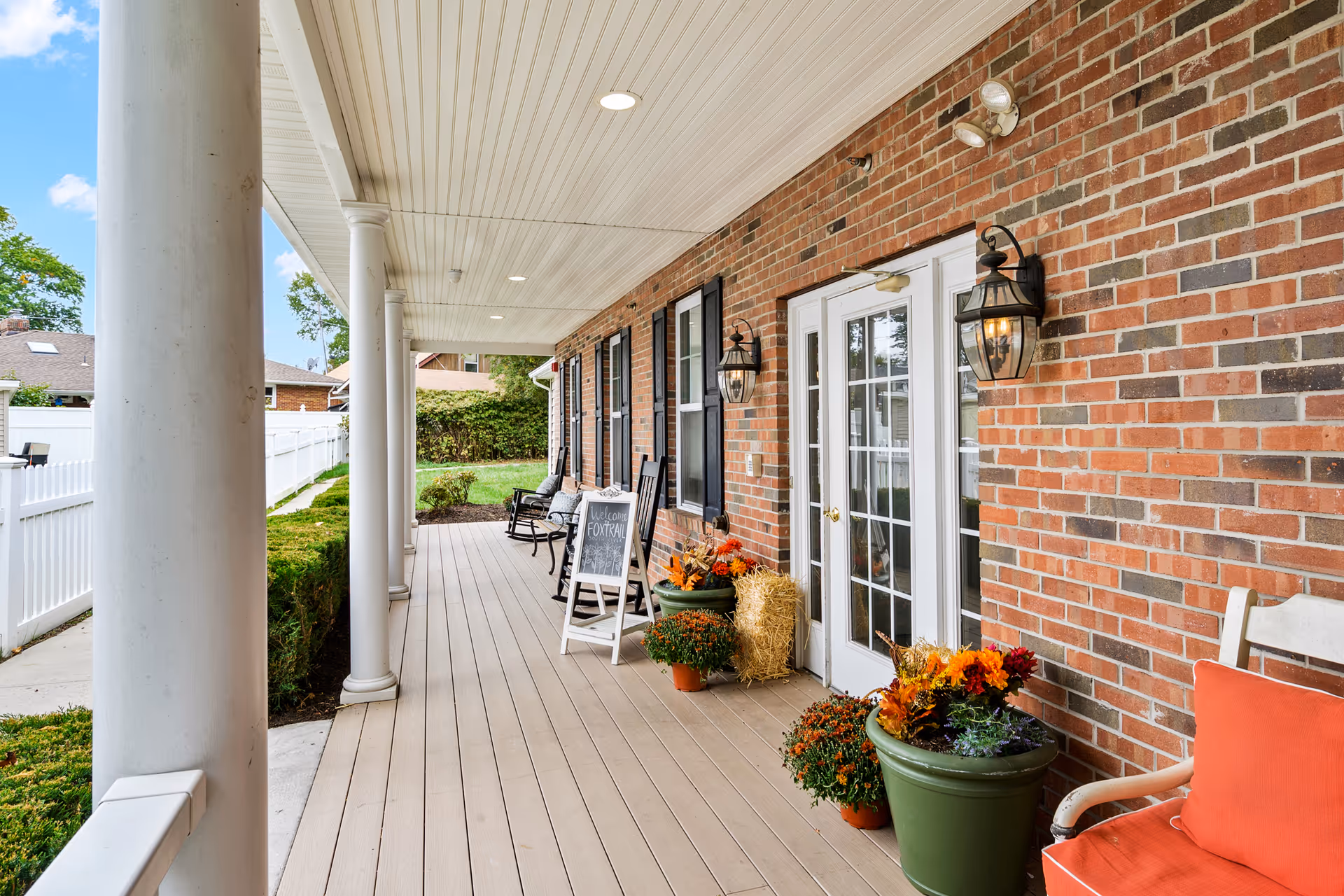 Covered porch area with white columns and a wooden floor at a brick building. The porch features black rocking chairs, potted plants with colorful flowers, a small hay bale, and a chalkboard sign that reads 'Welcome Fox Trail'. There are two wall-mounted lantern-style lights beside white French doors with glass panes. A white fence and greenery are visible along the side of the porch.