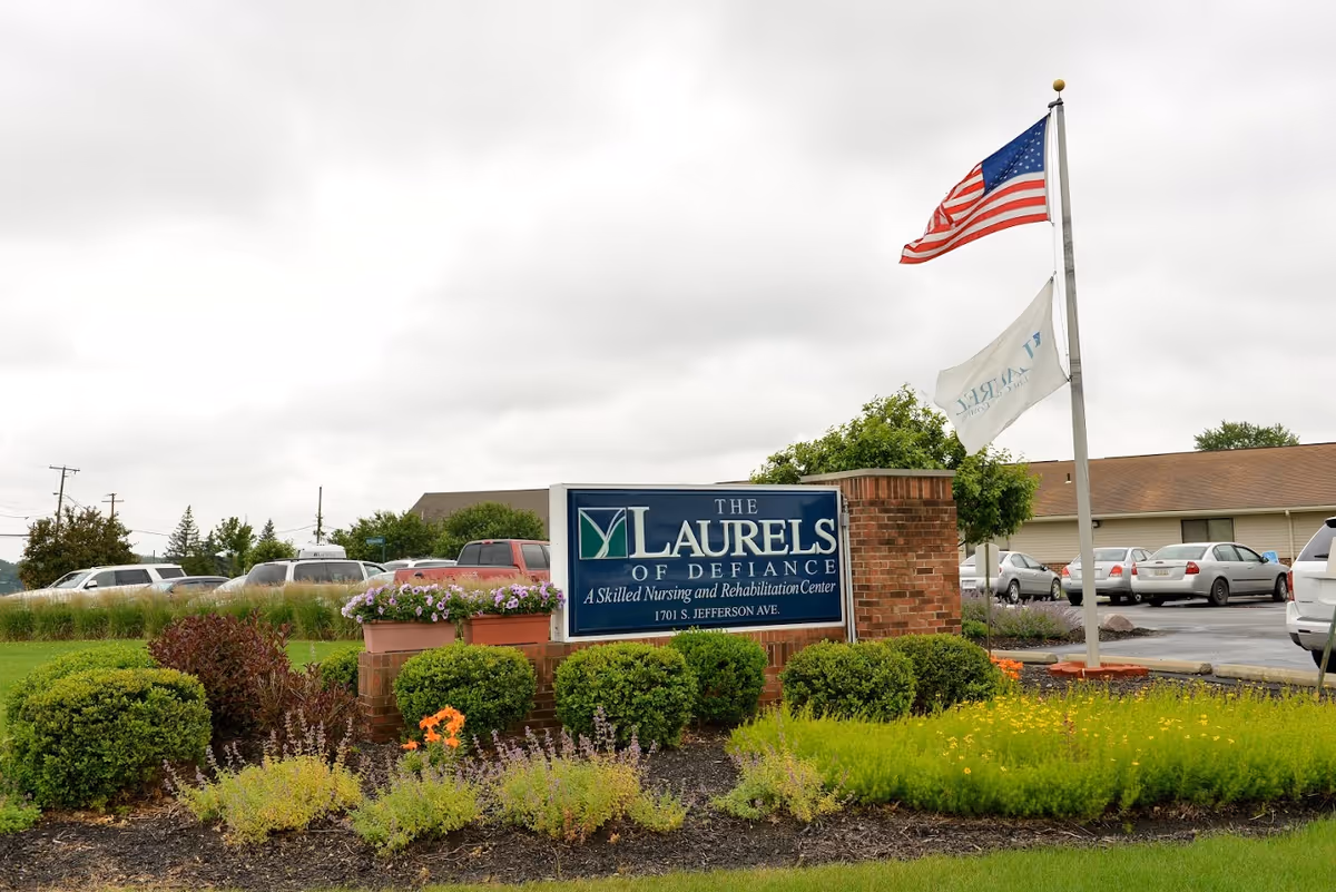 Sign for The Laurels of Defiance nursing and rehabilitation center in landscaped grounds with an American flag and parked cars behind it.