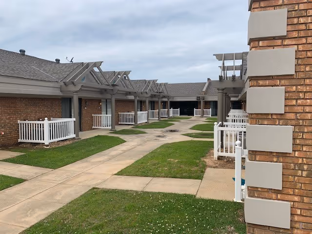 Outdoor courtyard area of The Gables At Spring Lake Assisted Living featuring single-story brick buildings with white railings, small patios, and a paved walkway surrounded by grass under a cloudy sky.