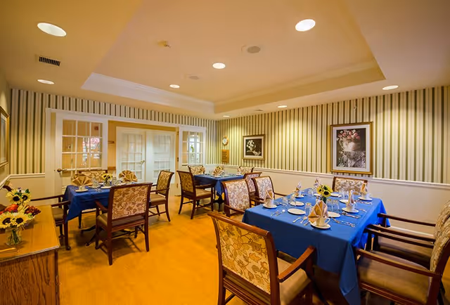 A dining room with several tables covered in blue tablecloths, each set with plates, napkins, and silverware. The room has striped wallpaper, framed floral artwork on the walls, and wooden chairs with floral upholstery. There is a sideboard with flower arrangements and double glass doors at the back.