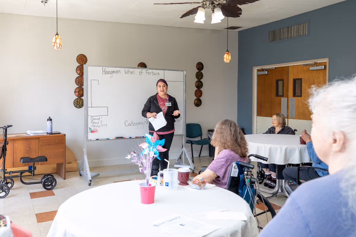 A group activity in a senior living facility room where a staff member stands in front of a whiteboard with the words 'Hangman Wheel of Fortune' written on it. Several elderly residents are seated around tables, some in wheelchairs, attentively watching the staff member. The room has light-colored walls, a ceiling fan, and pendant lights.