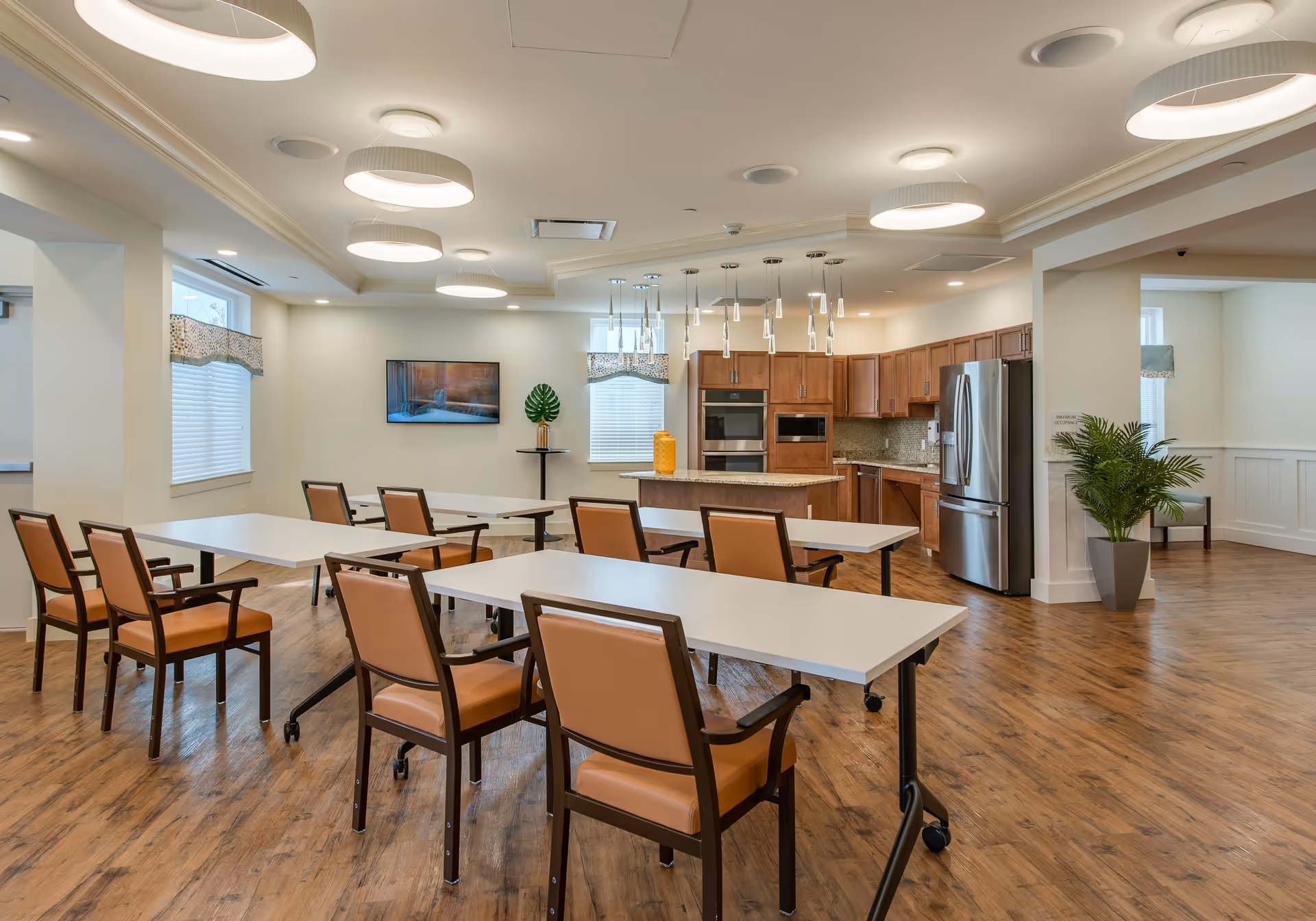 A spacious, well-lit room with several tables and chairs arranged in rows facing a kitchen area. The kitchen features wooden cabinets, a stainless steel refrigerator, built-in oven and microwave, and a central island with pendant lights hanging above. The room has wood flooring, large windows with blinds and valances, modern ceiling lights, and a flat-screen TV mounted on the wall. There are also decorative plants placed around the room.
