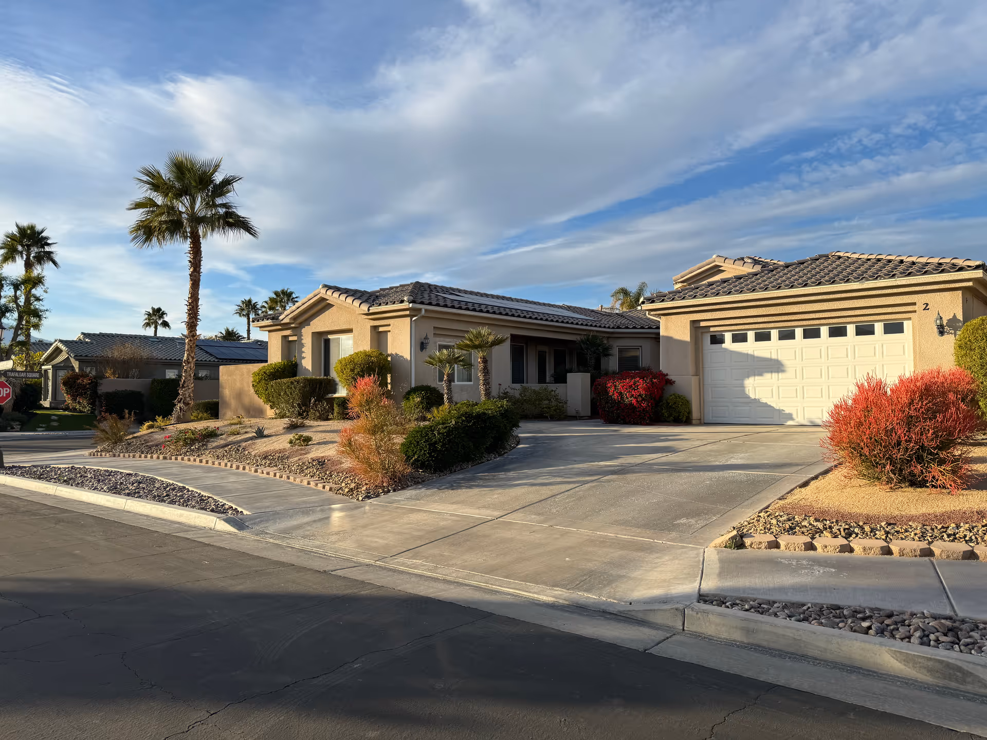 Exterior view of a single-story residential building with a tiled roof, a driveway leading to a white garage door, and landscaped front yard featuring palm trees, bushes, and decorative rocks under a partly cloudy sky.