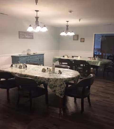 Communal dining room with two long tables covered in plastic floral tablecloths, dark chairs, overhead light fixtures, and a blue sideboard against the wall.