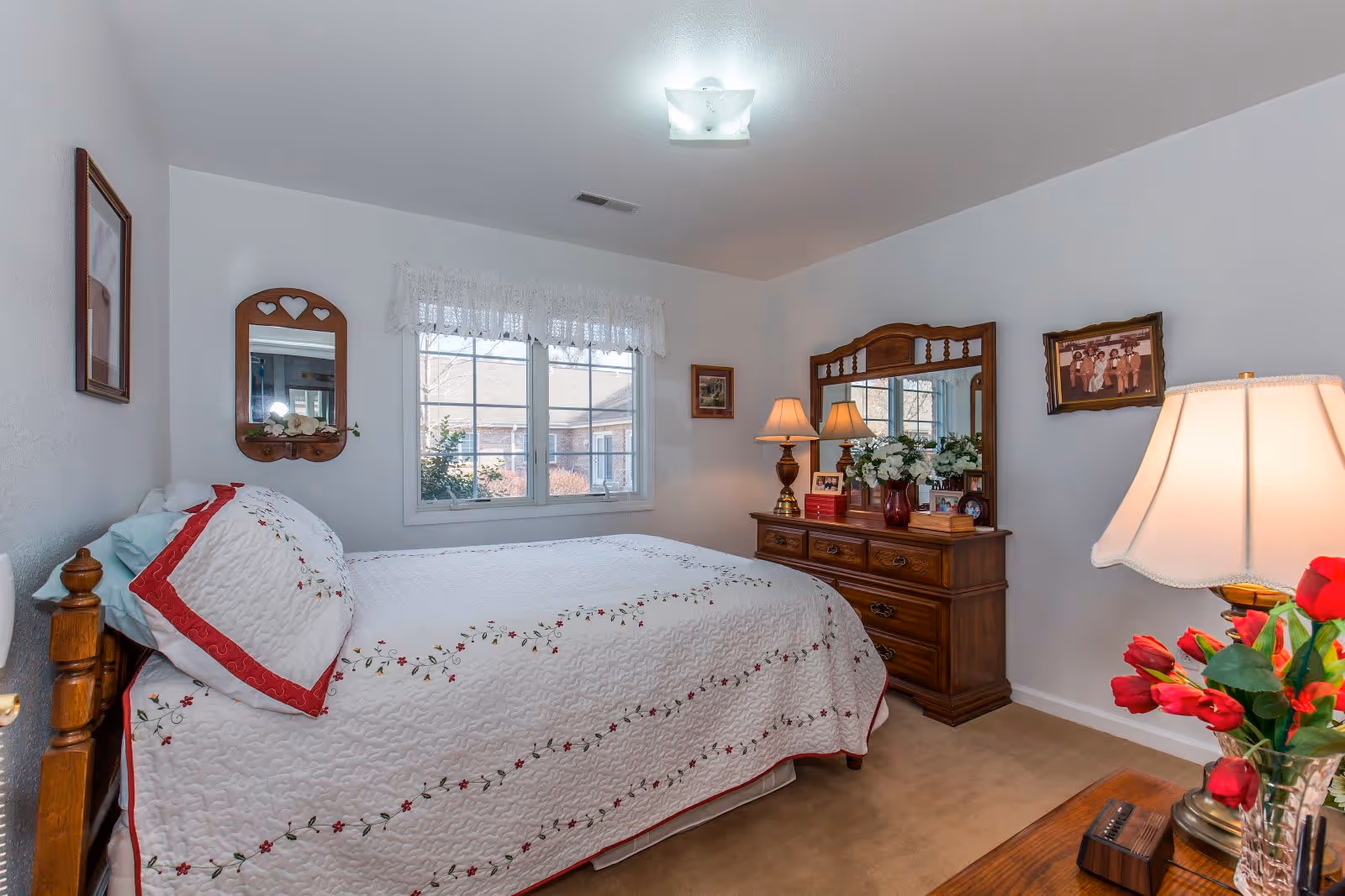 A cozy bedroom with a wooden bed frame and a white quilted bedspread decorated with red and green floral embroidery. The room has a window with white lace curtains, a wooden dresser with a large mirror, two table lamps, framed pictures on the walls, and a vase of red flowers on a wooden nightstand.