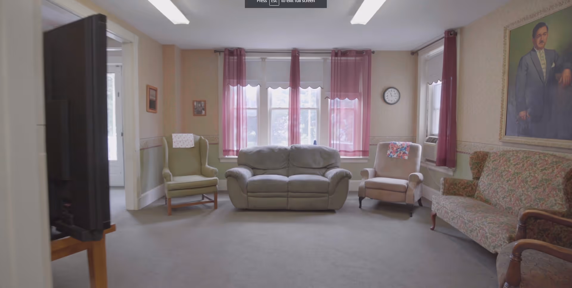 Sunlit common room with several upholstered chairs and a sofa arranged in front of windows with pink curtains.