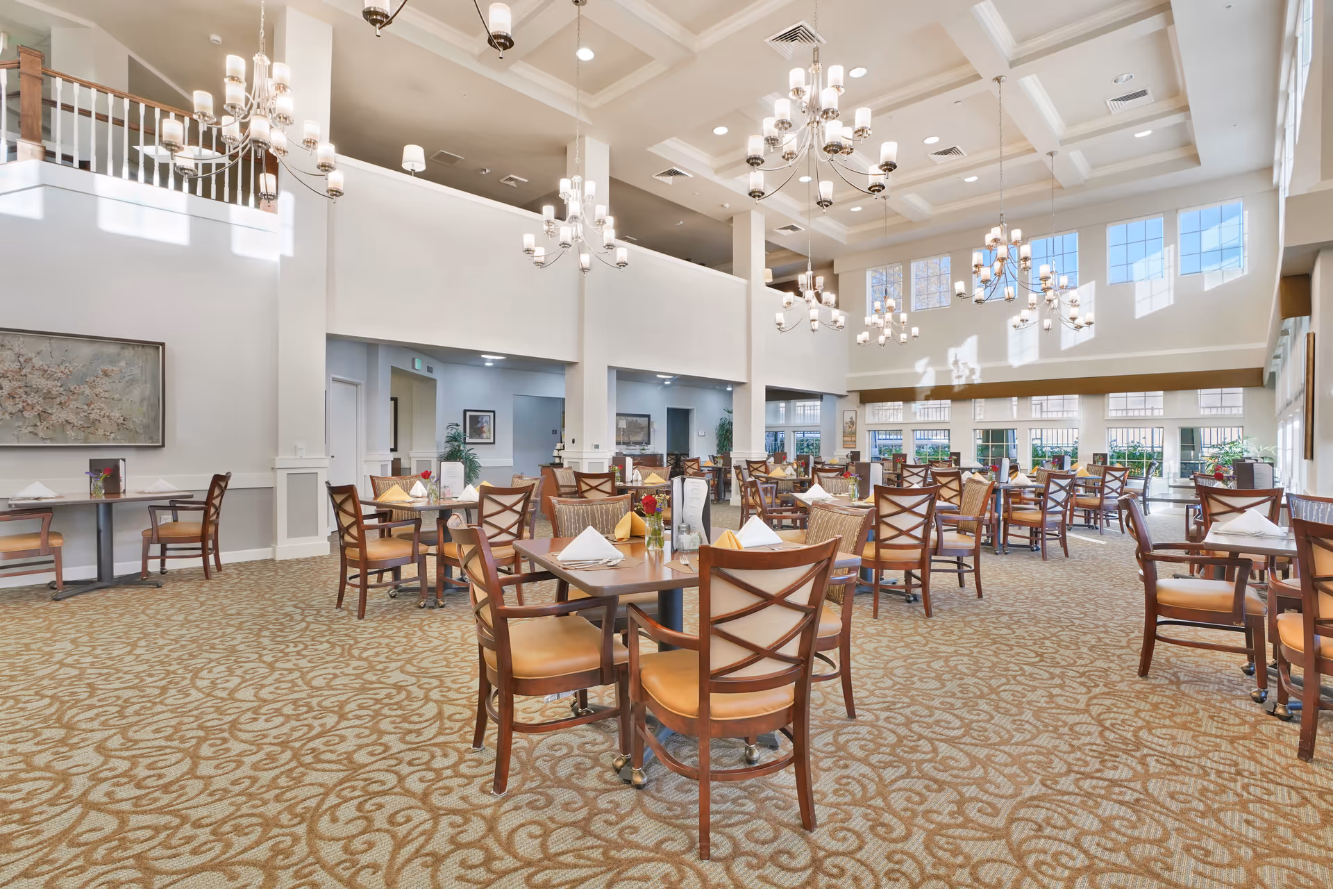 A spacious and well-lit dining room with multiple wooden tables and chairs arranged neatly. The room features high ceilings with several elegant chandeliers, large windows allowing natural light to fill the space, and a patterned carpet covering the floor. The tables are set with napkins and small decorative items, creating a welcoming atmosphere.