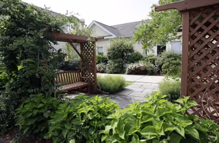 A peaceful garden area with lush green plants, a wooden swing bench under a wooden pergola, stone-paved walkway, and residential buildings in the background.