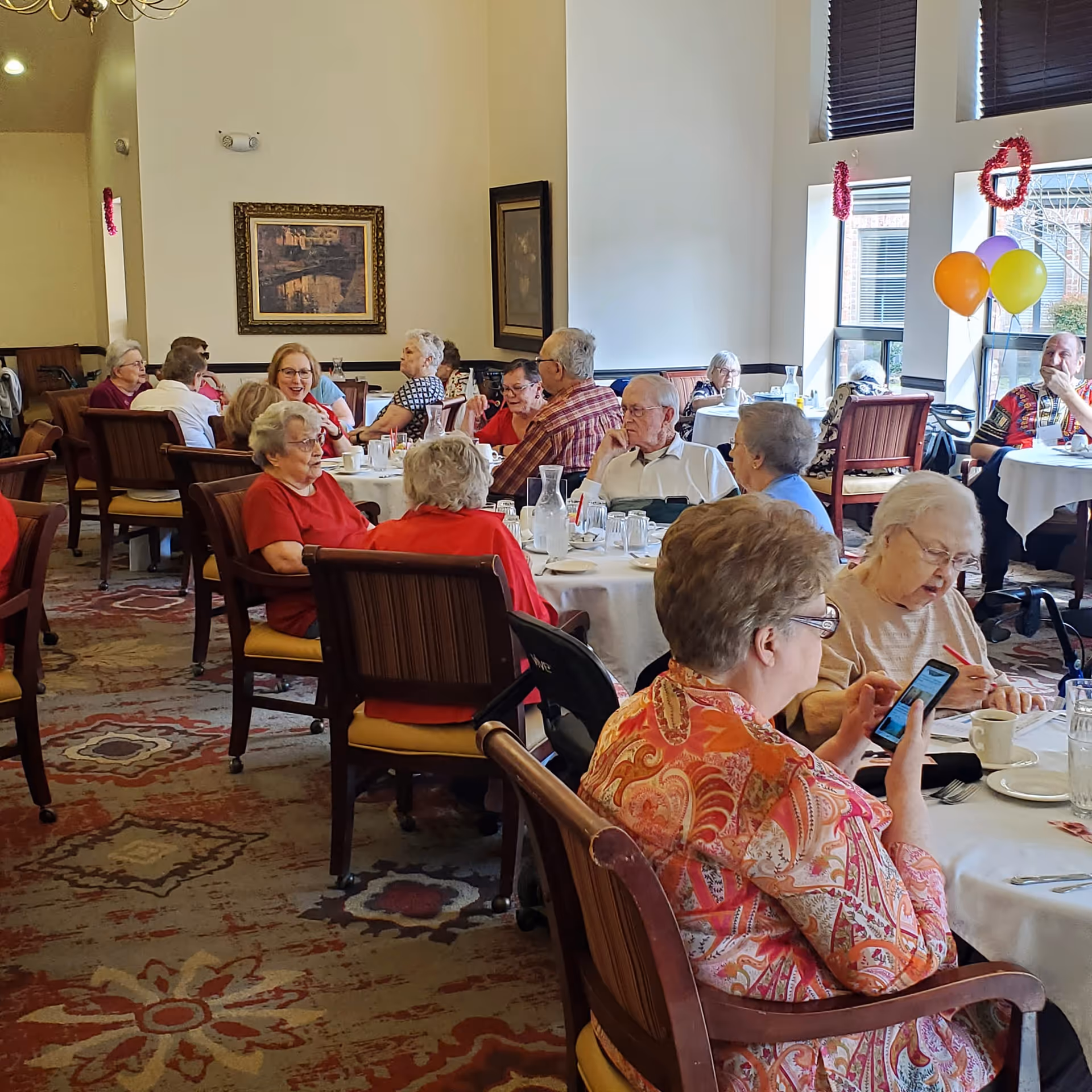 A group of elderly people sitting at round tables in a dining room, engaged in conversation and activities. The room has large windows with natural light, decorated with balloons and garlands. The tables are set with glasses, cups, and pitchers of water.