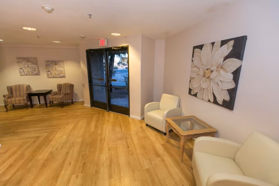 Lobby seating area with armchairs, small tables, wood-look flooring, and a large floral wall painting by glass double doors.