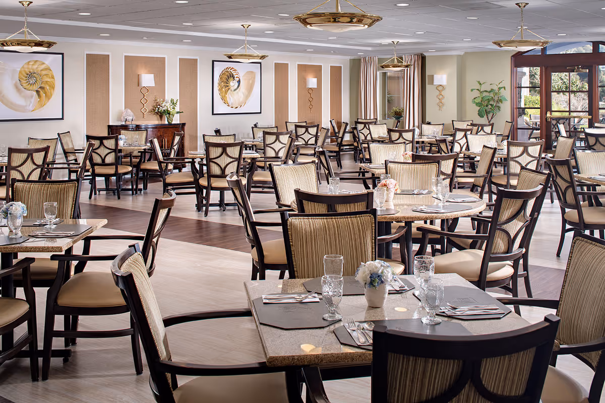 A spacious and well-lit dining room with multiple tables and chairs arranged neatly. Each table is set with placemats, glassware, and silverware, and some have small floral centerpieces. The room features large windows with curtains, decorative wall art, and elegant light fixtures hanging from the ceiling.