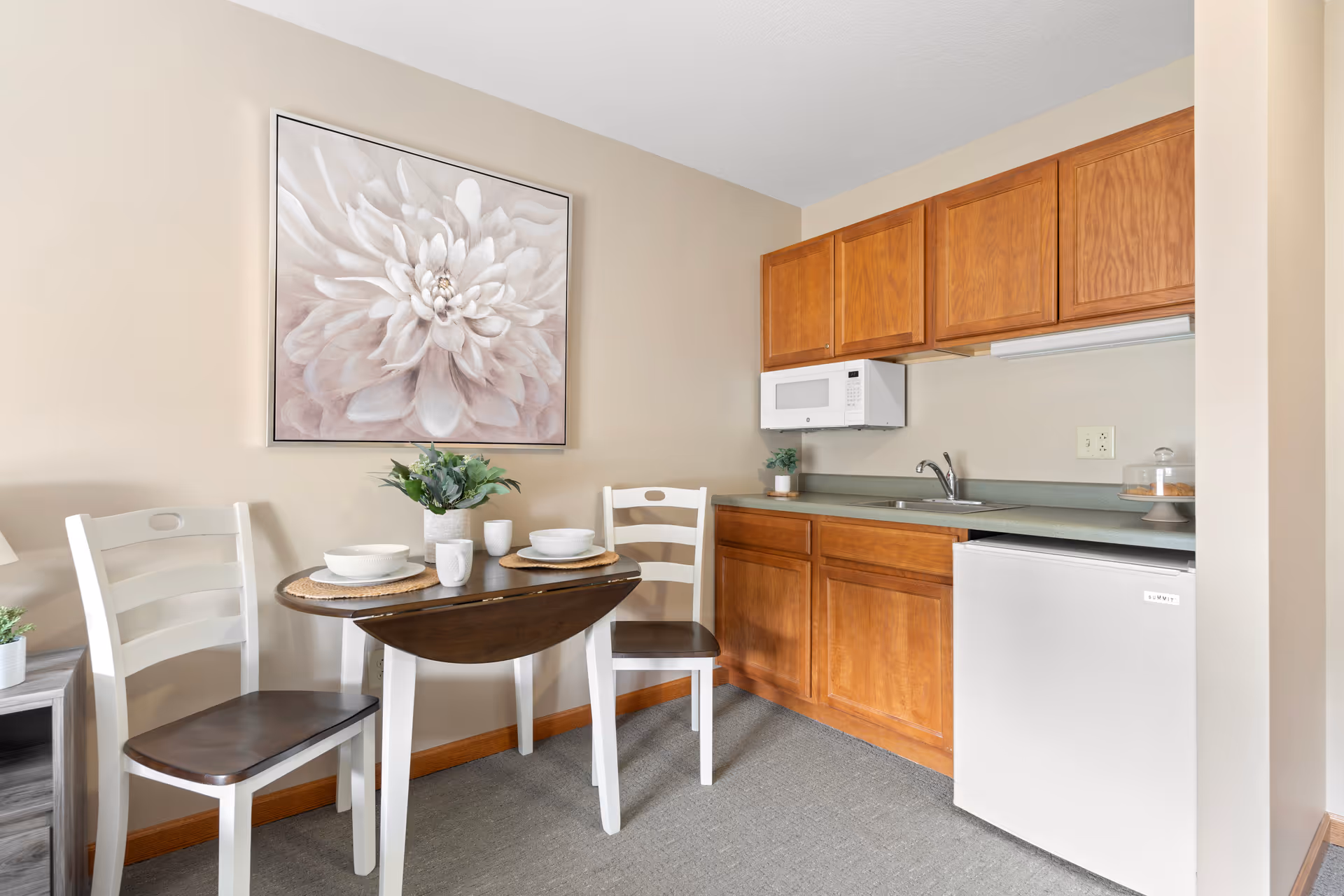 A small kitchenette area with wooden cabinets, a white microwave, a sink, and a mini refrigerator. In front of the kitchenette is a small wooden drop-leaf table set for two with white chairs, plates, cups, and a small plant. A large floral artwork hangs on the beige wall above the table.