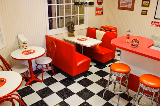 Retro diner-style dining area with red booths, round tables, and chrome counter stools on a black-and-white checkered floor.