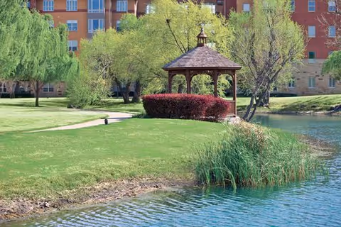 A peaceful outdoor scene at Highland Springs Senior Living Community featuring a wooden gazebo surrounded by green grass, trees, and shrubs next to a calm pond with clear blue water. In the background, a multi-story residential building is visible.