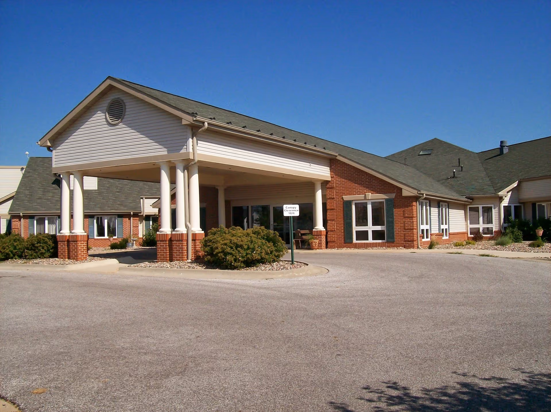Exterior front view of a single-story brick and siding building with a covered entrance supported by white columns, clear blue sky above, and a circular driveway in front.