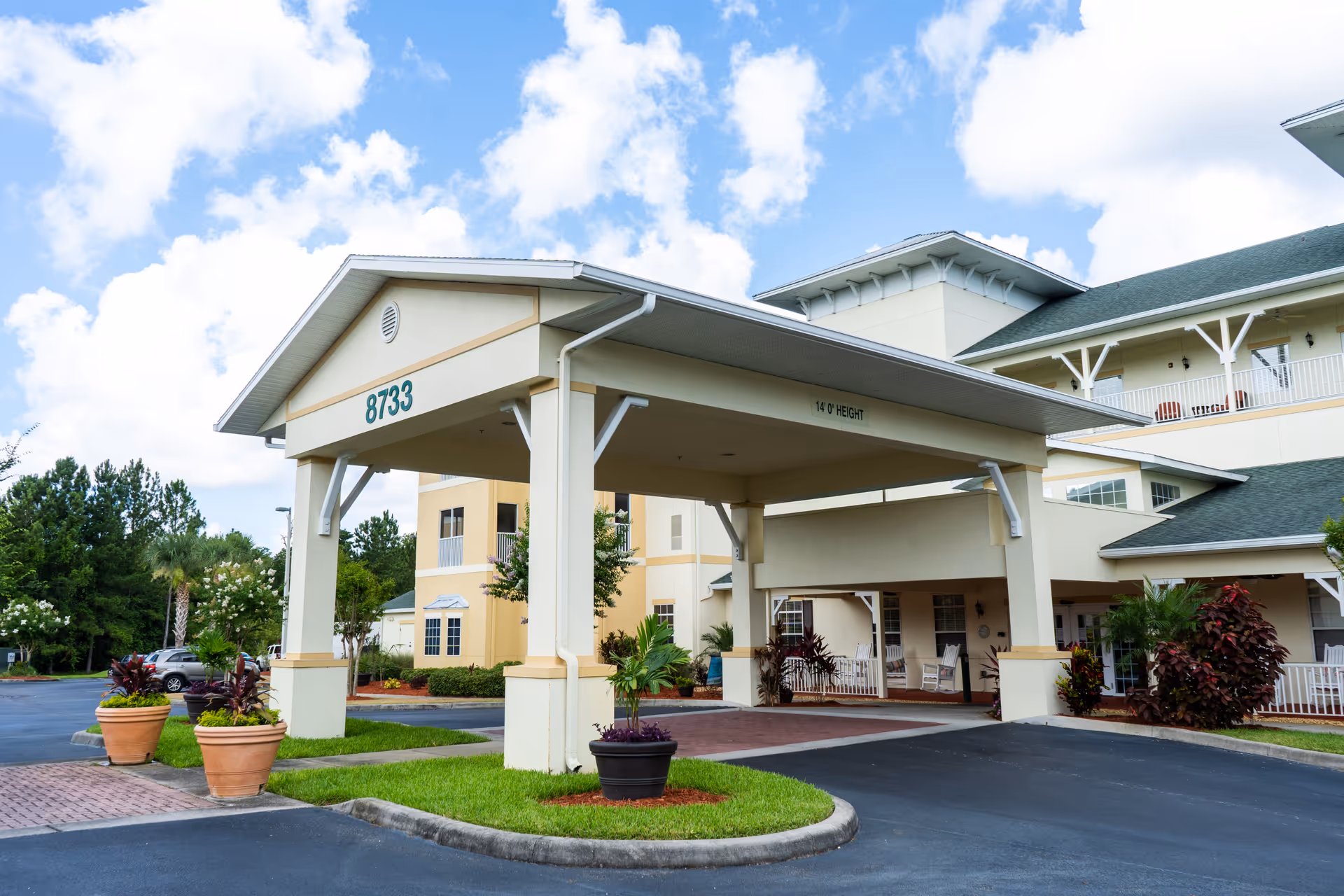 Entrance of a senior living facility named Sunflower Springs with a covered drop-off area marked with the number 8733. The building is light-colored with green roofing and surrounded by landscaped plants and trees under a partly cloudy sky.