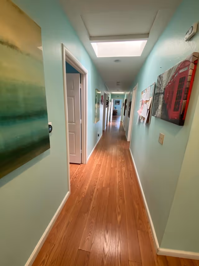 A long interior hallway with light teal walls, wood flooring, a skylight, and artwork and doors along the corridor.