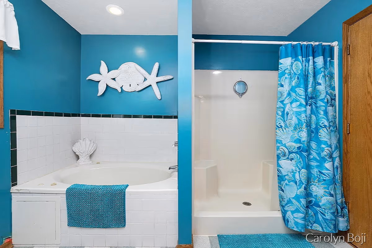 Bathroom with teal walls featuring a white corner bathtub with a teal towel hanging on the side, a white tiled surround, and a decorative shell sculpture. Next to the bathtub is a white shower stall with built-in seats and a blue shower curtain with a seashell pattern. A wooden door is visible on the right side.